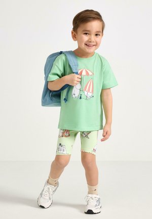 Giovane ragazzo sorridente che indossa una maglietta verde menta con stampa Moomin e pantaloncini, scarpe da ginnastica bianche, calzini beige e porta uno zaino blu su una spalla.