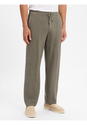 Uomo che indossa pantaloni in lino verde oliva con coulisse e scarpe beige senza lacci, in piedi davanti a uno sfondo bianco semplice.