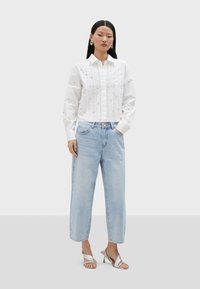 Chemise en coton blanche avec des broderies élaborées, manches longues et plis, associée à un jean en denim bleu clair, coupe droite, et des sandales à talons argentées.