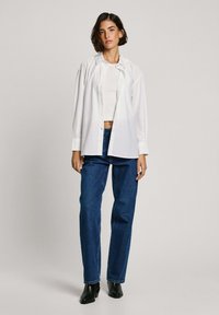 Weißes, button-up Hemd über einem taillierten weißen Oberteil, kombiniert mit blauen geraden Jeans und schwarzen Stiefeletten. Einfaches, lässiges Ensemble.
