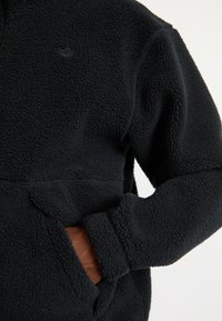 Pullover de forro polar negro con una superficie texturizada y peluda. Cuenta con un bolsillo frontal y un logotipo sutil en el pecho. Mangas largas y corte relajado.