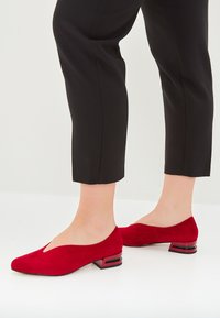 Rote Wildlederschuhe mit niedrigem Absatz und V-Ausschnitt-Design, ausgestattet mit einem kontrastierenden geometrischen Blockabsatz in Schwarz und Rot. Elegante und strukturierte Form.
