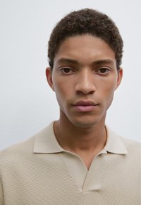 Beige texturerad polo-tröja med v-ringning och korta ärmar, bärs av en ung man med lockigt hår och en neutral bakgrund.