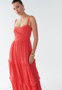 Robe maxi corail à volants, en tissu léger, avec bretelles ajustables, accents à volants et corsage cintré. Texture lisse sur toute la surface.