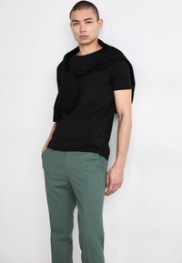 Zwarte t-shirt met korte mouwen en een ronde hals, gecombineerd met een slim-fit, groene textuurbroek; een zwarte trui over de schouders gedrapeerd.