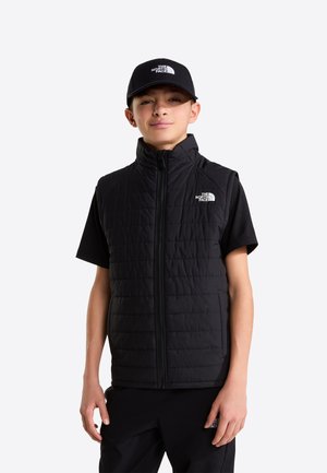 Niño con gorra negra de North Face, camiseta negra de manga corta y chaleco acolchado sin mangas negro, de pie frente a un fondo liso.