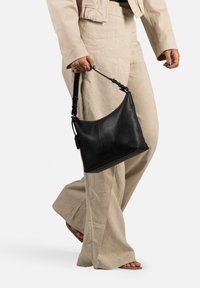 Borsa hobo in pelle nera con una texture morbida, tracolla regolabile e un piccolo tag con logo. Indossata in modo casual con pantaloni beige.