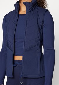 Námorně modrá sportovní bunda s prošívaným předním dílem, vysokým límcem a dvěma kapsami na zip po stranách. Vyrobena z hladkého, elastického materiálu.