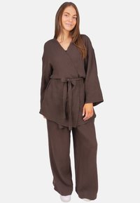 Frau steht lächelnd da und trägt eine locker sitzende braune Wickelbluse mit Bindegürtel und passende weit geschnittene Hosen vor weißem Hintergrund.