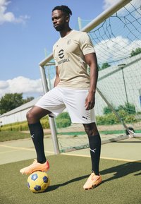 Beige fodboldtrøje med logo, hvide shorts, sorte knæhøje sokker og skrigorange fodboldstøvler. Bolden er gul med blå accenter.