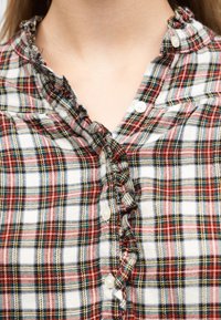 Κοντινό πλάνο πουκάμισου με καρό σχέδιο σε κόκκινο, λευκό, πράσινο και μαύρο, κουμπωμένο με βολάν κατά μήκος της γραμμής των κουμπιών και του περιλαίμιου.