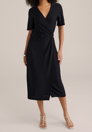 Kvinde iført en sort omslags-midi-kjole med korte ærmer og hvide hætte-sandaler med hæl, stående mod en ensfarvet beige baggrund.