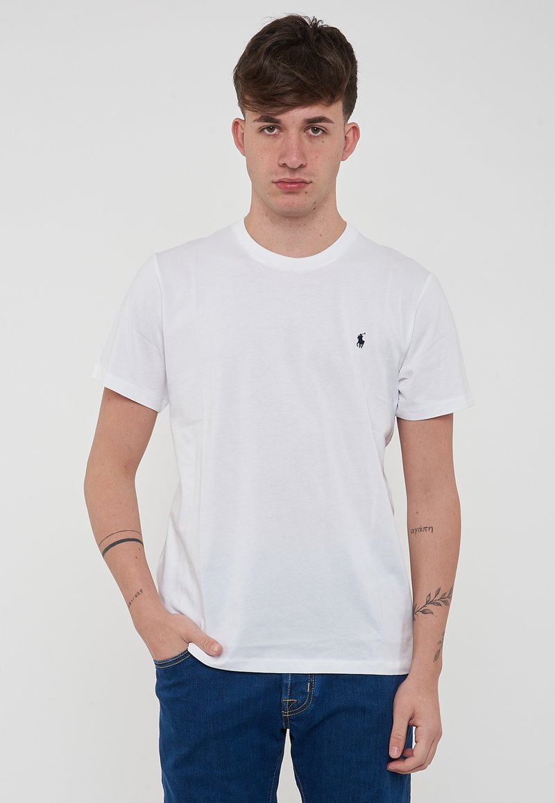 T-shirt bianco in cotone con colletto a girocollo e maniche corte, caratterizzato da un piccolo logo blu navy sul lato sinistro del petto. Indossato con jeans blu.