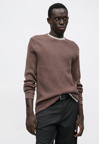 Brun ribstrikket sweater med rund halsudskæring og lange ærmer, lagdelt over en lysegrå skjorte; kombineret med mørkegrå bukser.