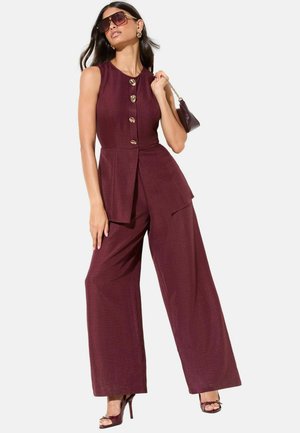 Vrouw draagt een mouwloze bordeaux jumpsuit met grote gouden knopen, draagt bijpassende tas, grote zonnebril en sandalen met hoge hakken.