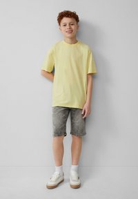 Camiseta de algodón amarilla, de mangas cortas, combinada con pantalones cortos de mezclilla grises. Zapatillas blancas con suela marrón y calcetines blancos completan el atuendo.