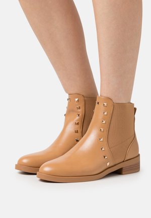 Stivali in pelle color cuoio alti fino alla caviglia, con borchie metalliche quadrate lungo i pannelli laterali elasticizzati, indossati su gambe nude su uno sfondo bianco.