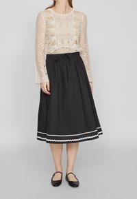 Lace-stickad krämfärgad långärmad topp med rundad halsringning, tillsammans med en svart ankellång kjol med vit vågig fåll och dragsko i midjan.