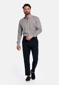 Man die loopt in een grijs geblokt overhemd met lange mouwen, zwarte broek en zwarte instappers tegen een effen witte achtergrond.