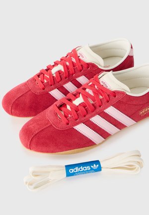 Para czerwonych zamszowych butów sportowych Adidas z różowymi paskami i czerwonymi sznurowadłami, oznaczonych "Tokyo,", pokazana z dodatkowymi białymi sznurowadłami Adidas.