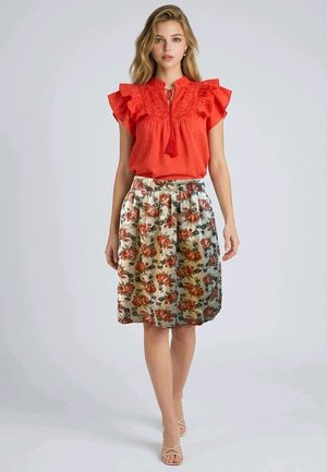 Mujer joven con blusa sin mangas roja con volantes y falda floral hasta la rodilla, de pie contra un fondo claro y liso.