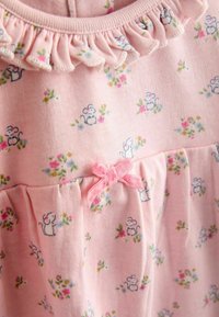 Rochie din bumbac roz cu un decolteu volanat, având un model floral și ilustrații cu șoareci mici. Accentuata cu o fundă roz la talie.