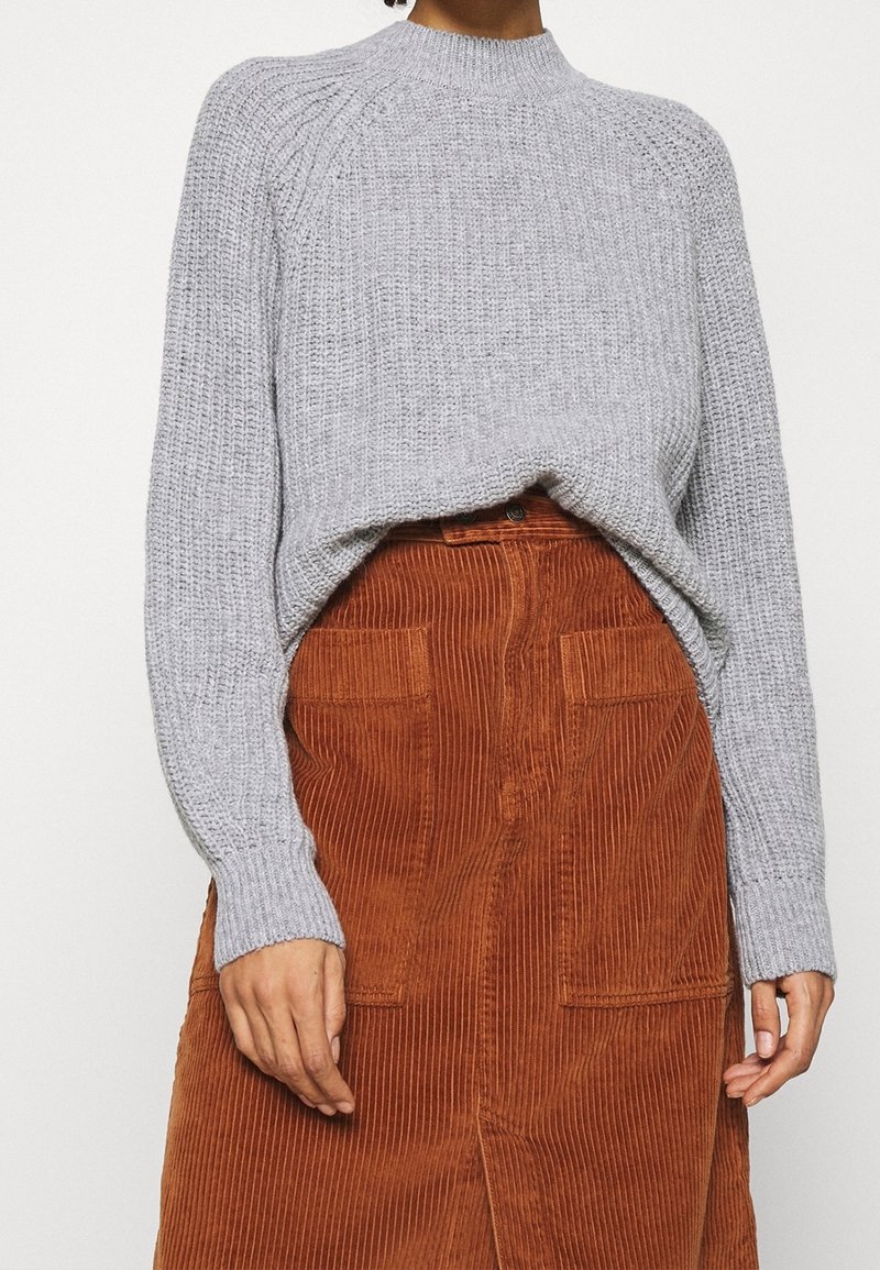 Femme portant un pull en maille gris clair rentré dans une jupe en velours côtelé orange rouille taille haute avec des poches plaquées à l'avant.