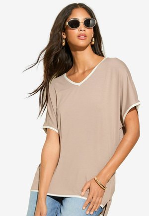 Beige losvallend T-shirt met een V-hals, witte rand langs de halslijn en mouwen, gecombineerd met lichtblauwe denim jeans.