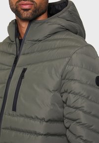 Olivgrön vadderad jacka med huva, med horisontell sömnad, två fickor med dragkedja och en rund logga på axeln.