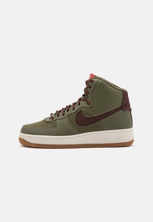 Zapatilla alta con un upper sintético de color verde oliva, acentos de cuero marrón, cordones rojos y una suela de goma blanca con una suela exterior de goma texturizada.