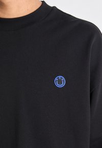 Felpa nera a girocollo con una piccola toppa circolare blu che presenta una faccina sorridente e le lettere "HGO" sul lato sinistro del petto.
