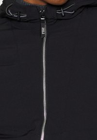 Sort lynlåsjakke med aftagelig hætte, der har en metallisk lynlås, elastiske snore og en glat stoftekstur.