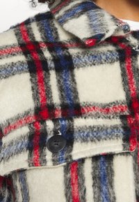 Ruudullinen fleece-takki, jossa on vaaleancreminvärinen pohja ja punaisia, sinisiä sekä mustia viivoja. Huomionarvoisia yksityiskohtia ovat musta nappi ja käännettävä kaulus.