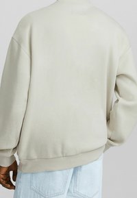 Persona che indossa un maglione beige oversize e jeans di denim azzurro chiaro, vista da dietro con una mano rilassata e l'altra piegata.