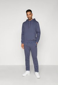 Navy hoodie with a front pocket, paired with matching navy jogger pants. Made of smooth fabric, features white sneakers and a logo tag.