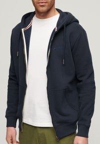 Granatowa bluza z kapturem na suwak, z kieszeniami kangurkami, regulowanym kapturem i haftowanym logo. Nałożona na białą koszulkę, zaprezentowana na neutralnym tle.