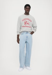 Grå sweatshirt med rød "ICECREAM SKATEBOARDS" tekst, løstsiddende lyseblå jeans og hvide sneakers. Modellen står foran en ensfarvet baggrund.