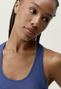 Tamsiai mėlynos spalvos sportinė marškinėlė, pasižyminti tekstūruota medžiaga su briaunotais raštais ir mažomis perforacijomis petnešėlėse.