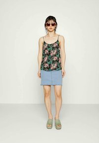 Top de tanque preto com padrão floral e alças finas, combinado com uma saia de ganga clara; modelo usando sapatos de plataforma verdes e óculos de sol. Fundo simples.