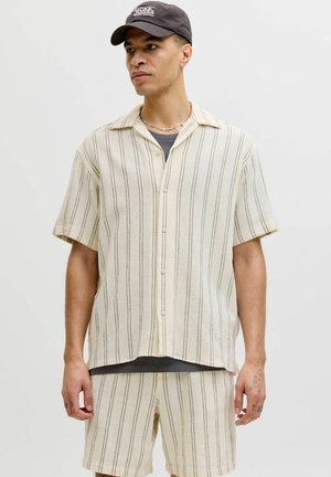 Homme portant une chemise beige à manches courtes et à boutons avec des rayures verticales fines, un short assorti, une casquette foncée et un collier chaîne en argent, fond neutre.