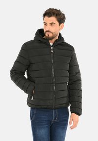 Uomo che indossa una giacca nera trapuntata con cappuccio e zip frontale, mani nelle tasche, abbinata a jeans blu, mentre guarda di lato.