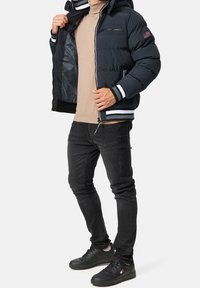 Musta toppatakki hupulla, jossa on kontrastivalkoiset raidat hihojen resoreissa ja helmassa, vetoketjulliset taskut ja teksturoitu materiaali. Päällä ruskeansävyisen paidan ja farkkujen kanssa.