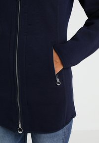 Merensininen vetoketjullinen takki, joka on valmistettu kuvioidusta kankaasta ja jossa on sivutaskut hopeanvärisillä vetoketjullisilla yksityiskohdilla sekä hieman kaareva helma.