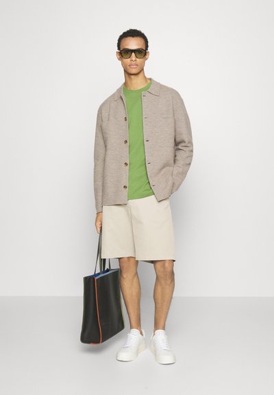 Cămașă călduță de culoare bej cu nasturi peste un tricou verde, pantaloni scurți bej, adidași albi și o geantă de umăr neagră cu accente colorate și curele.