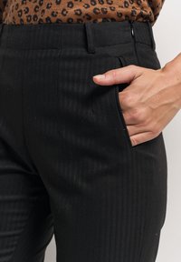 Pantalons à rayures noires en tissu texturé ; dotés d'une poche latérale et d'une taille ajustée. Le design comprend une fermeture à glissière.