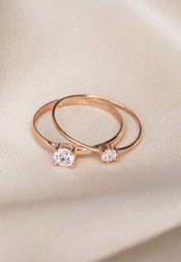 Twee rosegouden ringen, één met een grote ronde steen en één kleinere, tegen een zachte beige stof achtergrond. Vloeiend oppervlak, minimalistisch ontwerp.