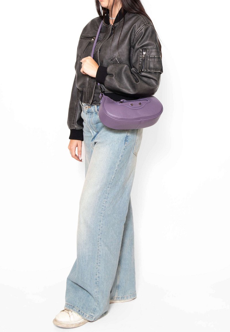 Borsa a tracolla in pelle viola con una forma curva, dotata di una cerniera superiore e di una tracolla removibile, abbinata a una giacca bomber nera e jeans chiari.