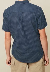 Camicia a maniche corte in tessuto blu scuro con un motivo geometrico testurizzato. Presenta un colletto casual e un orlo arrotondato, con un dettaglio sulla spalletta posteriore.