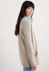 Beige adīta kardigāna ar atvērtu priekšpusi, garām piedurknēm un divām priekšējām kabatām. Iezīmējas ar ribotām malām un mīkstu tekstūru, uzvilkta virs zila krekla.