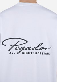 Camiseta de algodón blanca que presenta un gran logo en cursiva negra que dice "Pegador" con "TODOS LOS DERECHOS RESERVADOS" debajo.